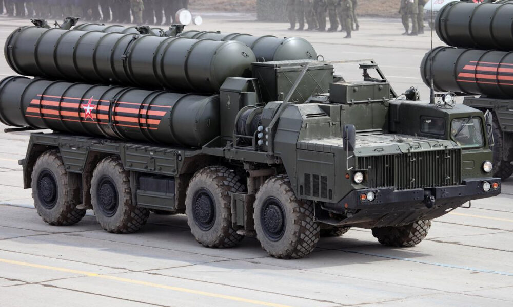 Eight-wheeled military transporter truck carrying large cylindrical missiles on a flatbed, olive drab with red star markings on the side crane area behind; troops visible in background.