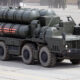 Eight-wheeled military transporter truck carrying large cylindrical missiles on a flatbed, olive drab with red star markings on the side crane area behind; troops visible in background.