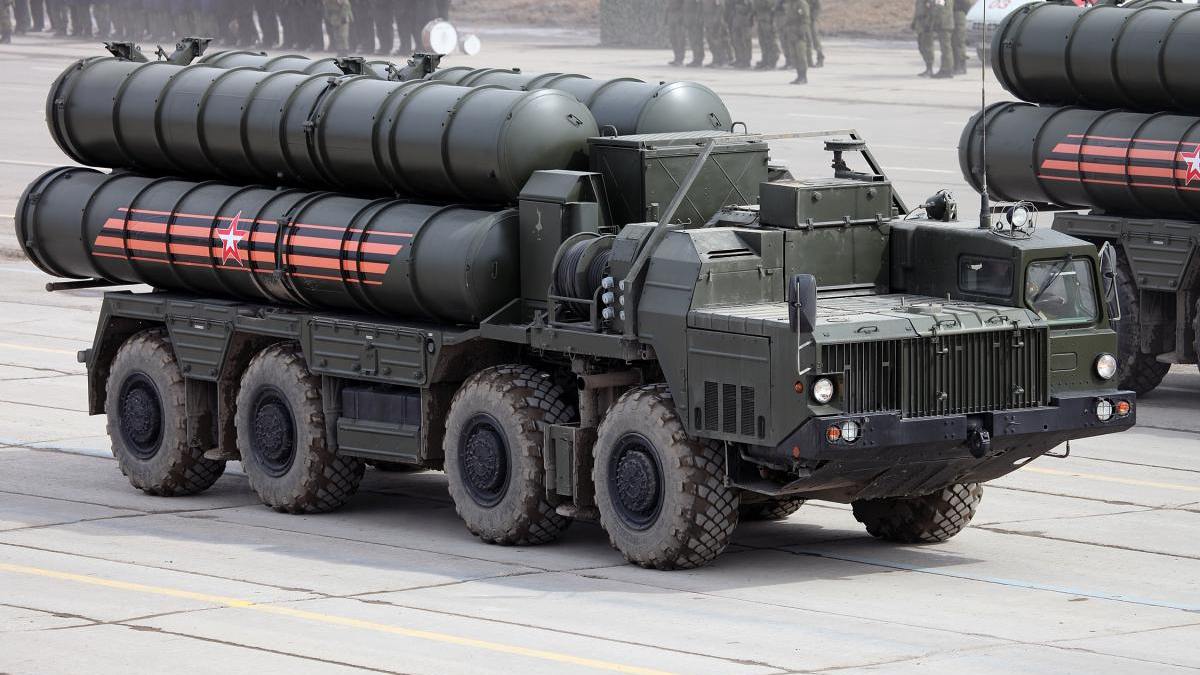 Eight-wheeled military transporter truck carrying large cylindrical missiles on a flatbed, olive drab with red star markings on the side crane area behind; troops visible in background.