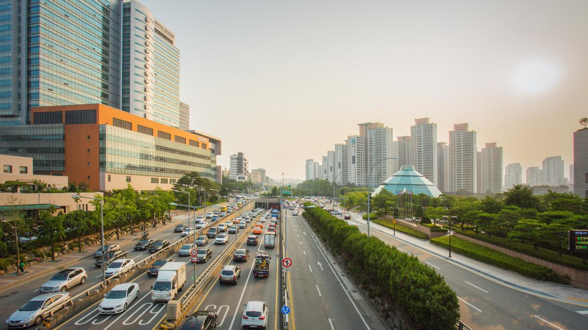 Busy urban highway lined with modern skyscrapers and a glass pyramid building at sunset, with heavy traffic on both sides.