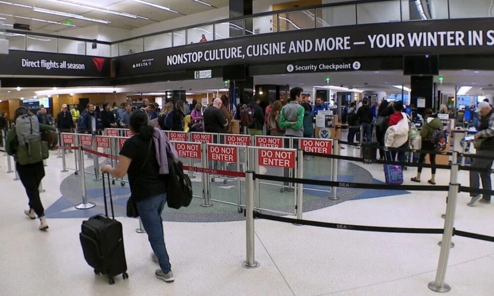 Busy airport security checkpoint with a long queue behind red 'Do Not Enter' barriers; passengers with rolling luggage wait as overhead signage advertises nonstop culture and travel.