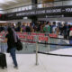 Busy airport security checkpoint with a long queue behind red 'Do Not Enter' barriers; passengers with rolling luggage wait as overhead signage advertises nonstop culture and travel.