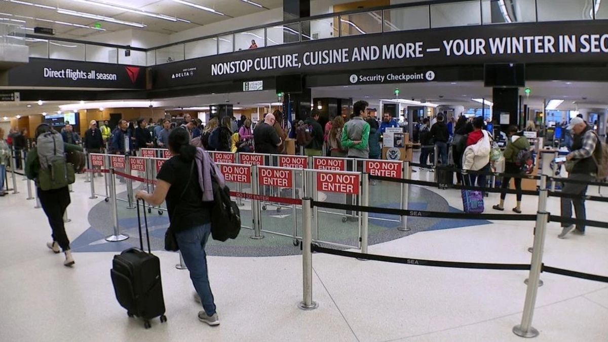 Busy airport security checkpoint with a long queue behind red 'Do Not Enter' barriers; passengers with rolling luggage wait as overhead signage advertises nonstop culture and travel.