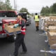 A volunteer in a red vest loads a cardboard box from a red pickup truck at a food drive, with stacked donation boxes nearby.
