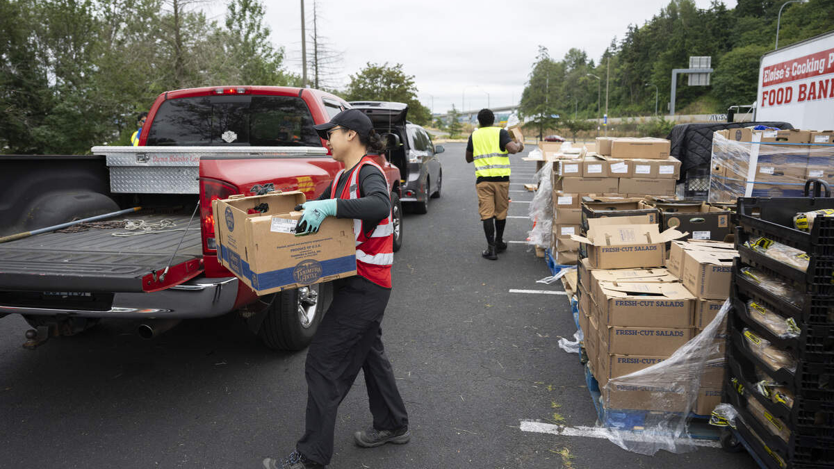 A volunteer in a red vest loads a cardboard box from a red pickup truck at a food drive, with stacked donation boxes nearby.
