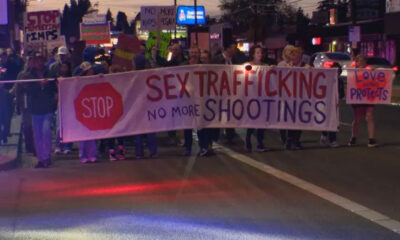 Group of protesters marching at night, carrying a large banner that says 'Sex Trafficking, No More Shootings' with a red stop sign on the left.