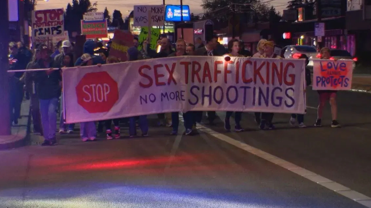 Group of protesters marching at night, carrying a large banner that says 'Sex Trafficking, No More Shootings' with a red stop sign on the left.