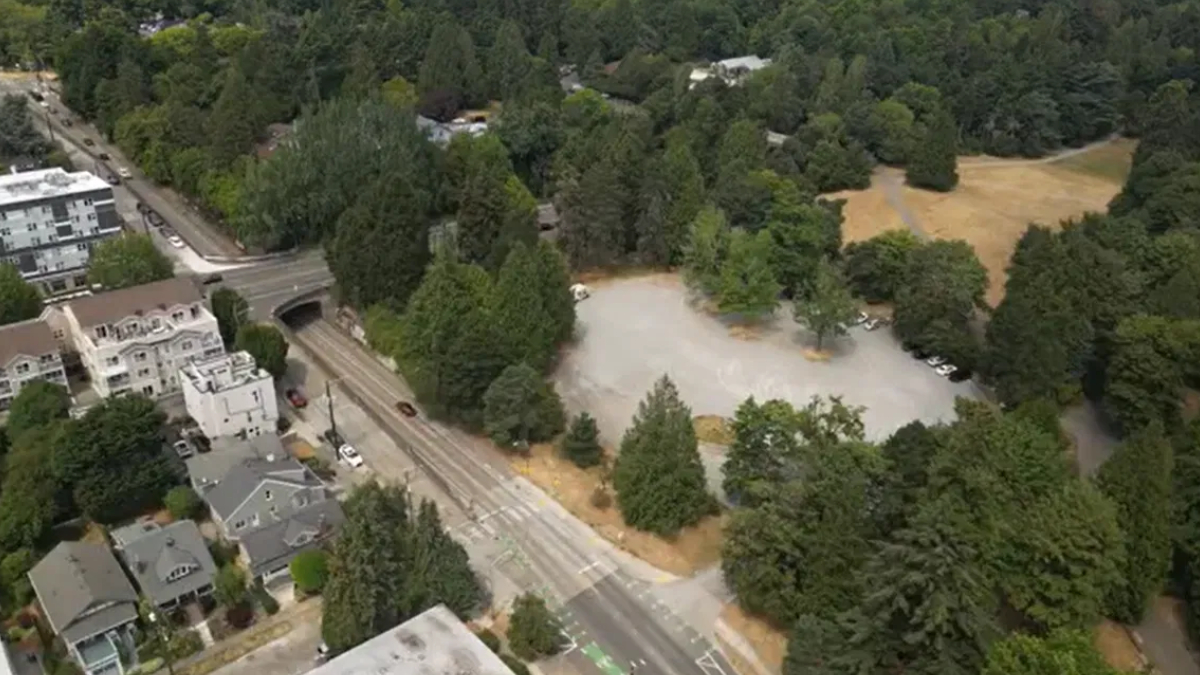 Aerial view of a street with residential buildings on the left, a railroad, and a large open gravel parking lot surrounded by trees.