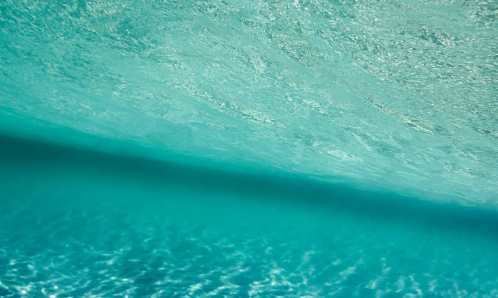 Aerial view of turquoise ocean water with sunlight ripples and a dark diagonal shadow across the surface