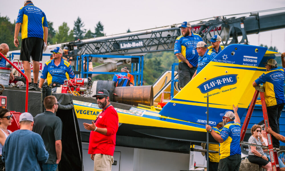 Crew members in blue and yellow uniforms work on a bright blue-and-yellow race boat on a trailer at a race event, with logos visible like Flav-R-Pac.