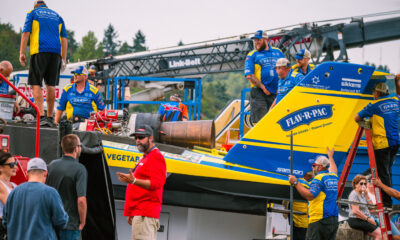 Crew members in blue and yellow uniforms work on a bright blue-and-yellow race boat on a trailer at a race event, with logos visible like Flav-R-Pac.