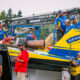 Crew members in blue and yellow uniforms work on a bright blue-and-yellow race boat on a trailer at a race event, with logos visible like Flav-R-Pac.