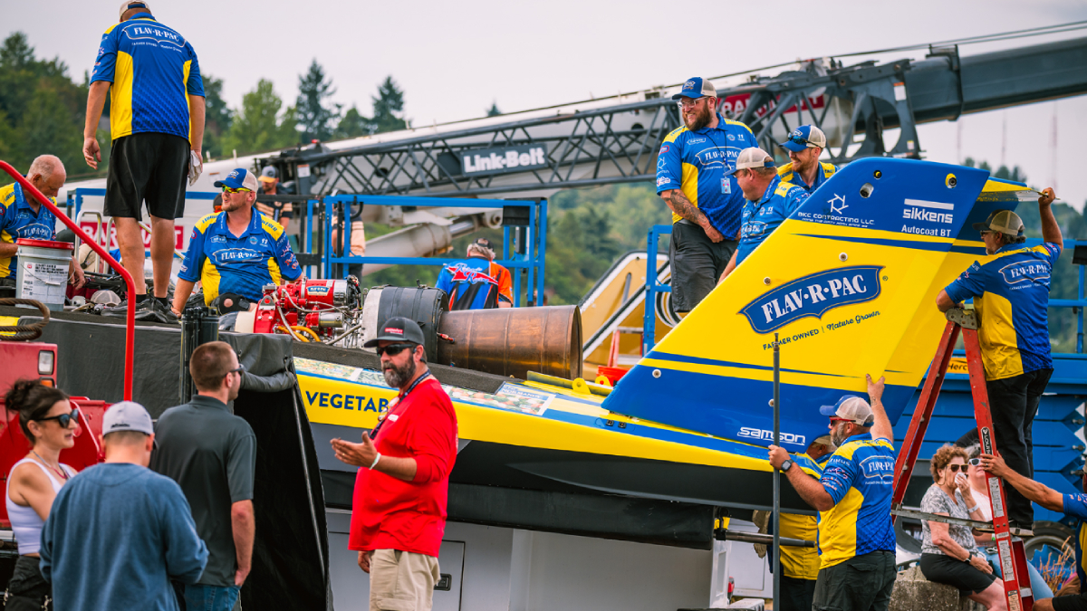 Crew members in blue and yellow uniforms work on a bright blue-and-yellow race boat on a trailer at a race event, with logos visible like Flav-R-Pac.