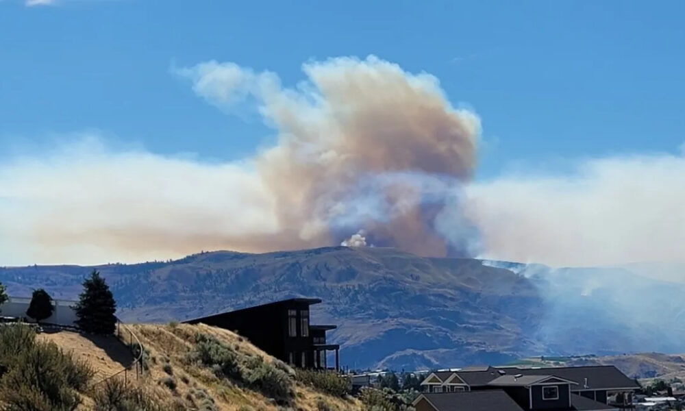 Smoke plume rising above distant hills behind a suburban neighborhood, under a blue sky, indicating a wildfire nearby.