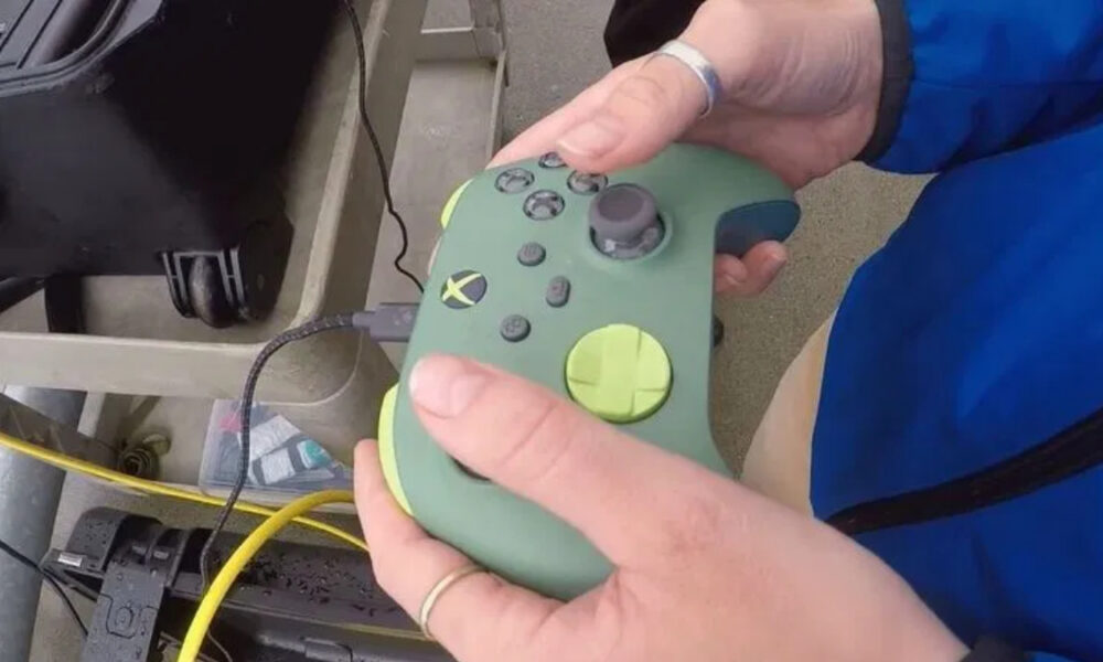 Person holds a green handheld controller with multiple buttons and a large joystick-like stick, connected by a cable to equipment nearby.