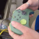 Person holds a green handheld controller with multiple buttons and a large joystick-like stick, connected by a cable to equipment nearby.