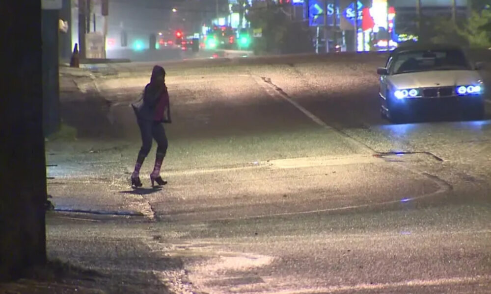 Person crossing a wet city street at night as a car with blue headlights approaches from the right.