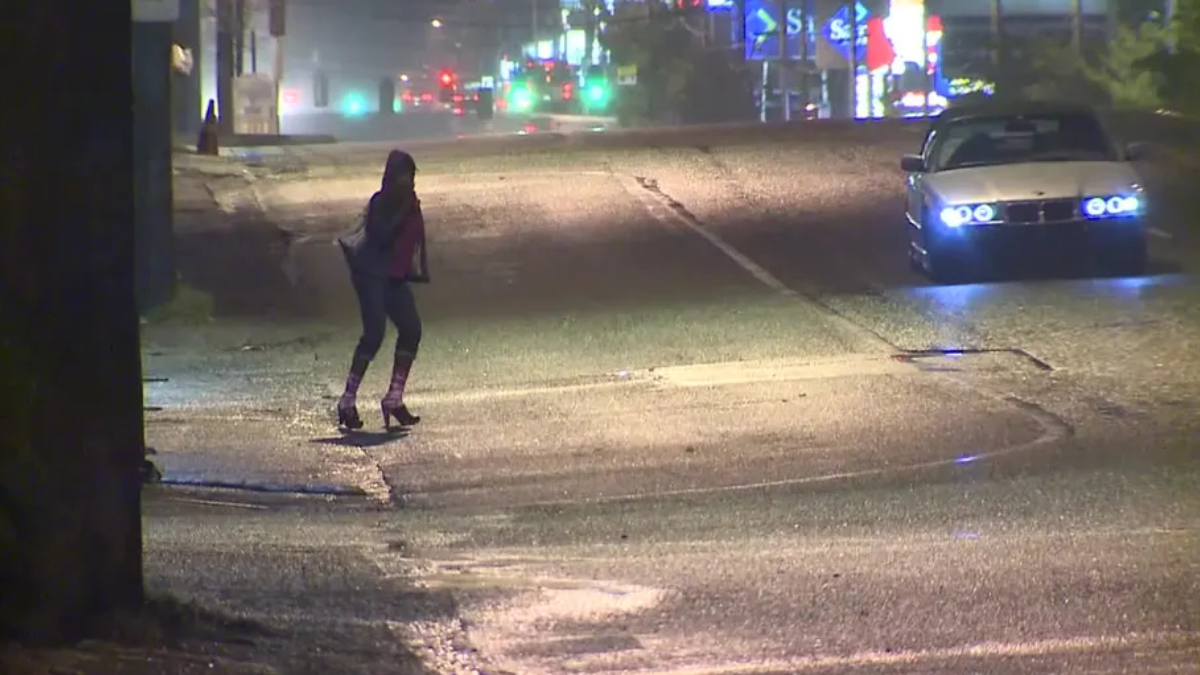 Person crossing a wet city street at night as a car with blue headlights approaches from the right.