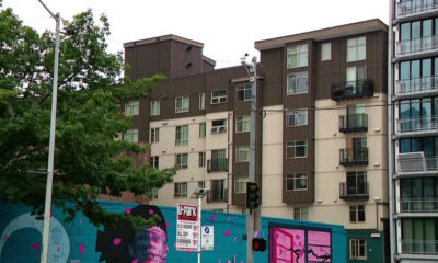 Urban street scene with a colorful mural on a blue wall in front of beige and brown apartment buildings; a green tree on the left.