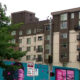 Urban street scene with a colorful mural on a blue wall in front of beige and brown apartment buildings; a green tree on the left.