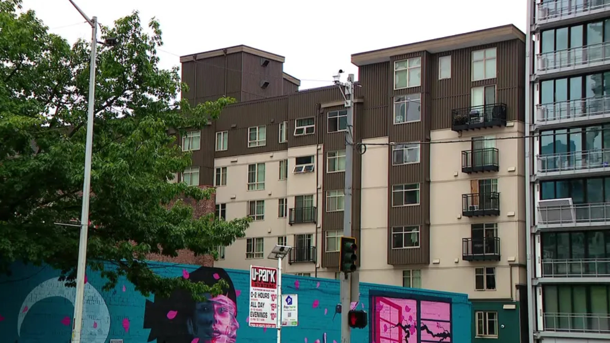 Urban street scene with a colorful mural on a blue wall in front of beige and brown apartment buildings; a green tree on the left.