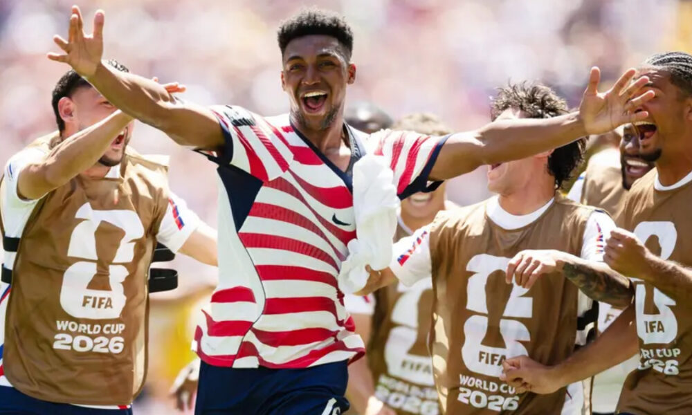 Soccer players in World Cup 2026 bibs celebrate with arms raised and smiles on a sunny field.