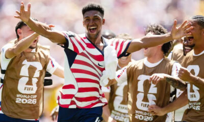 Soccer players in World Cup 2026 bibs celebrate with arms raised and smiles on a sunny field.