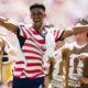 Soccer players in World Cup 2026 bibs celebrate with arms raised and smiles on a sunny field.