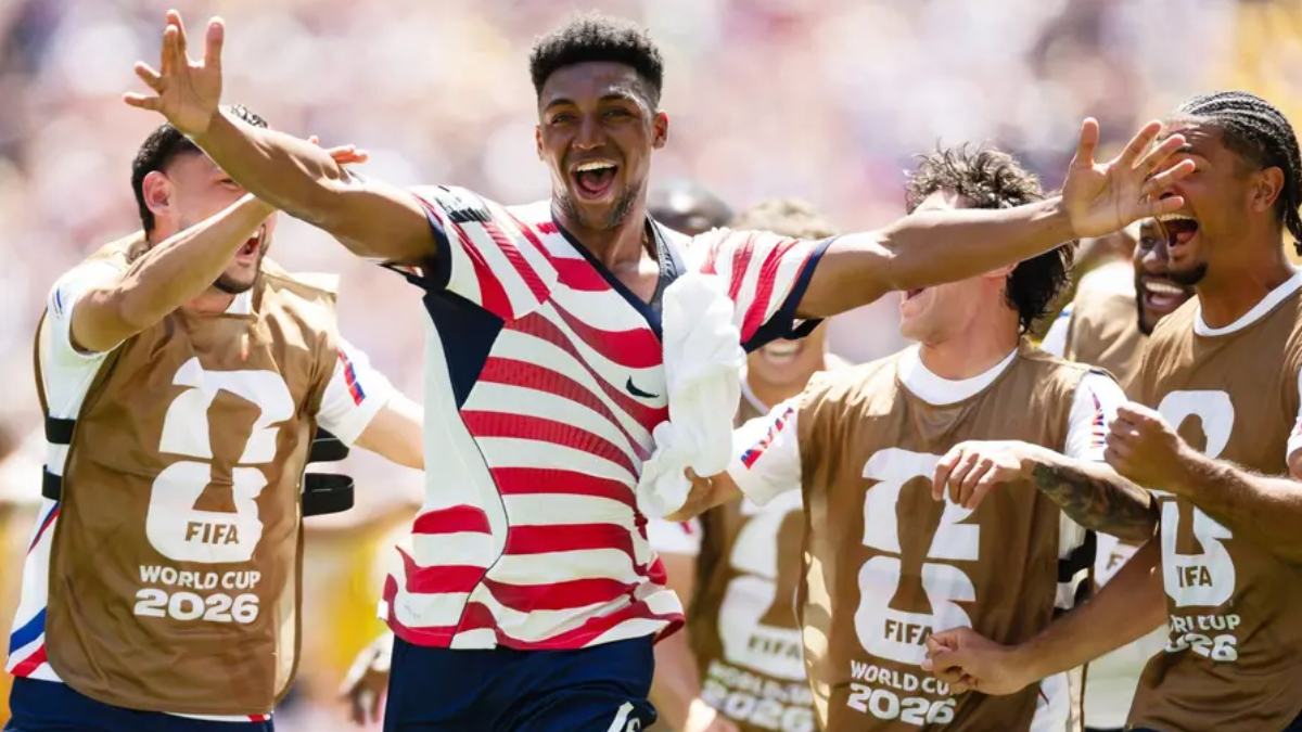 Soccer players in World Cup 2026 bibs celebrate with arms raised and smiles on a sunny field.