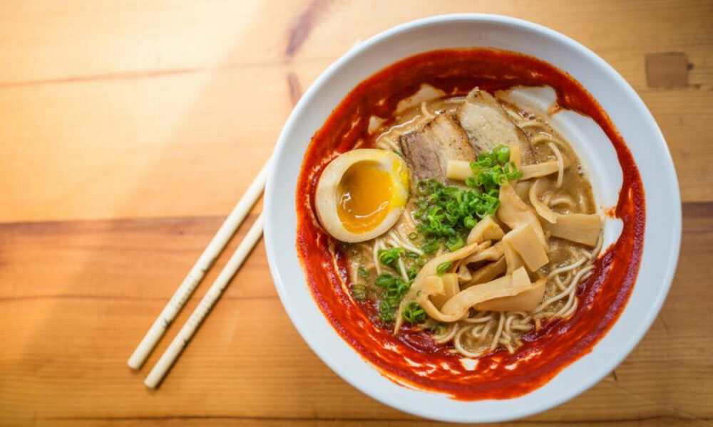 Spicy ramen in a white bowl with a soft-boiled egg, sliced pork, bamboo shoots, and green onions; chopsticks beside on a wooden table.