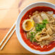 Spicy ramen in a white bowl with a soft-boiled egg, sliced pork, bamboo shoots, and green onions; chopsticks beside on a wooden table.