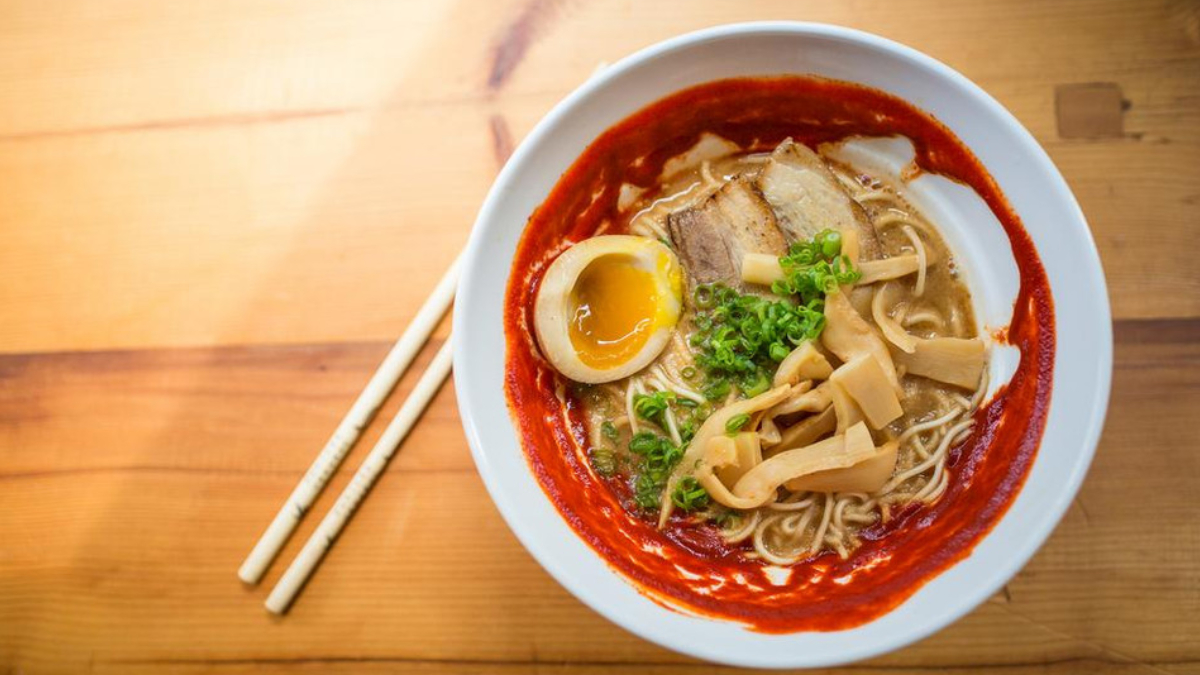 Spicy ramen in a white bowl with a soft-boiled egg, sliced pork, bamboo shoots, and green onions; chopsticks beside on a wooden table.