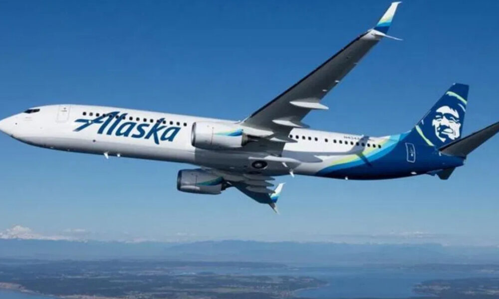 Airplane in-flight with Alaska Airlines livery, white fuselage and blue tail featuring a stylized face logo, flying over a landscape below