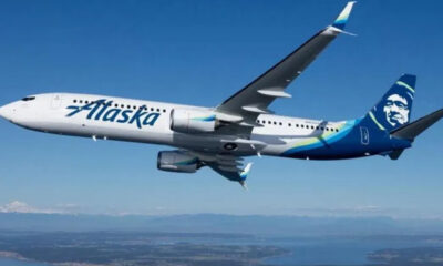 Airplane in-flight with Alaska Airlines livery, white fuselage and blue tail featuring a stylized face logo, flying over a landscape below