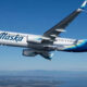 Airplane in-flight with Alaska Airlines livery, white fuselage and blue tail featuring a stylized face logo, flying over a landscape below