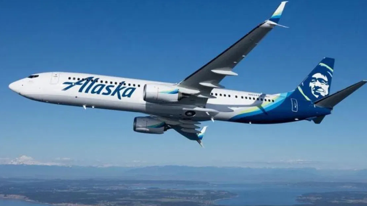 Airplane in-flight with Alaska Airlines livery, white fuselage and blue tail featuring a stylized face logo, flying over a landscape below