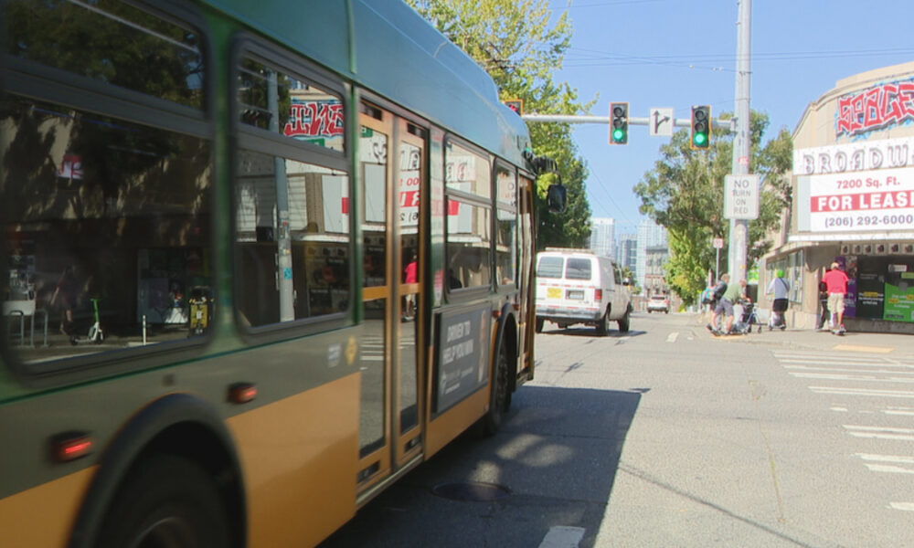 Green-and-yellow city bus dominates foreground as it stops on a street; pedestrians near the crosswalk and storefronts with a large 'FOR LEASE' sign in the background.