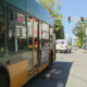 Green-and-yellow city bus dominates foreground as it stops on a street; pedestrians near the crosswalk and storefronts with a large 'FOR LEASE' sign in the background.