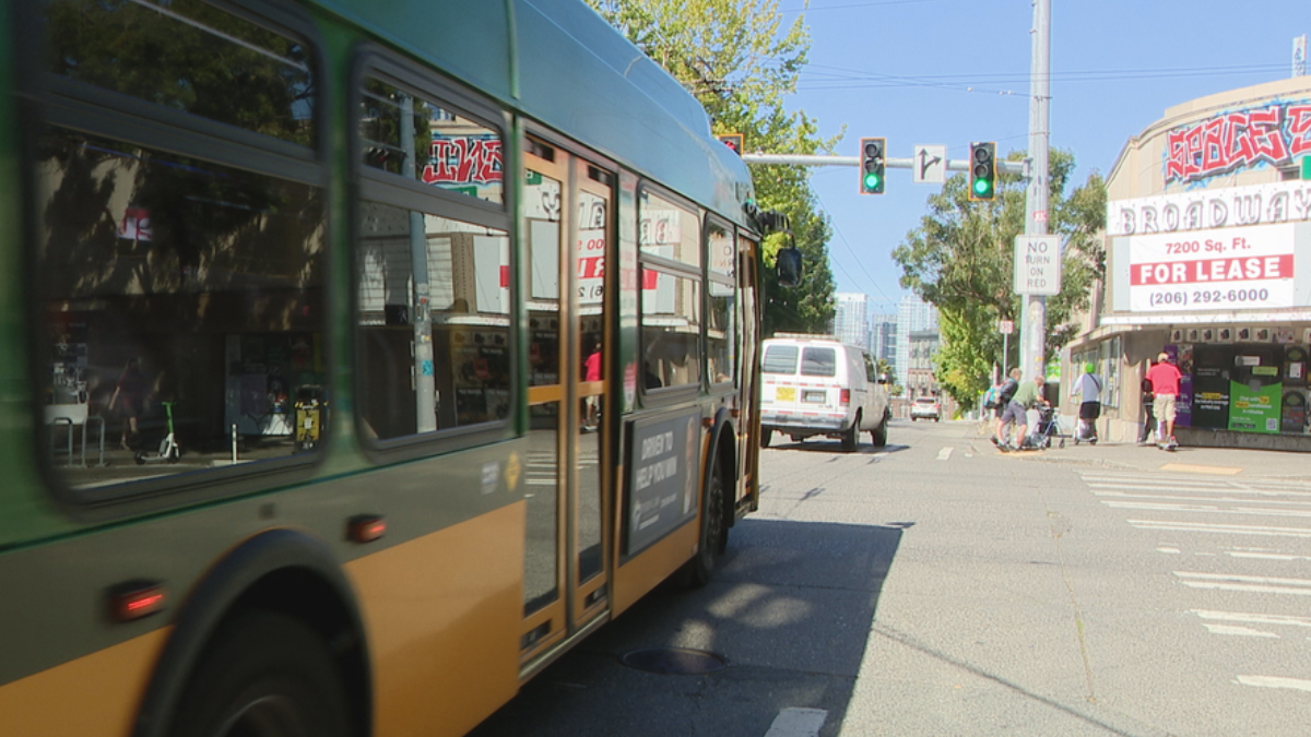 Green-and-yellow city bus dominates foreground as it stops on a street; pedestrians near the crosswalk and storefronts with a large 'FOR LEASE' sign in the background.