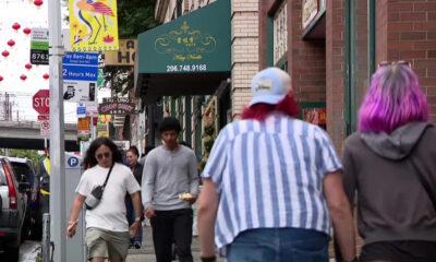 Busy city sidewalk with pedestrians and storefronts, red lantern decorations overhead.