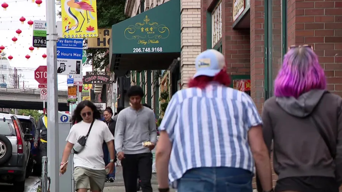 Busy city sidewalk with pedestrians and storefronts, red lantern decorations overhead.