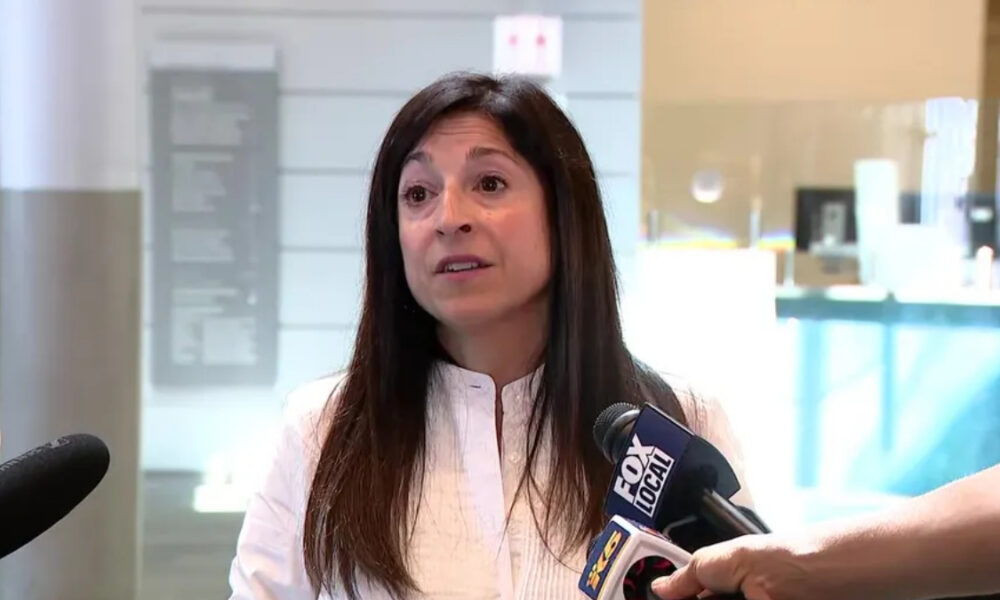 Woman in a white blouse being interviewed by reporters in a bright indoor lobby, microphones held close to her face.