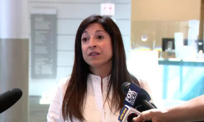 Woman in a white blouse being interviewed by reporters in a bright indoor lobby, microphones held close to her face.