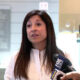 Woman in a white blouse being interviewed by reporters in a bright indoor lobby, microphones held close to her face.