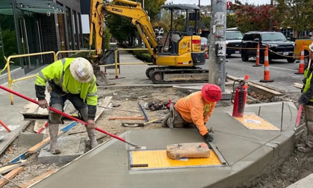 Construction workers in hi-visibility gear smooth fresh concrete along a curb on a city sidewalk尋