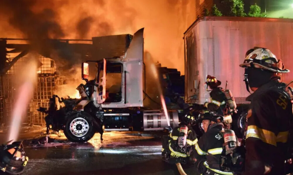 Crew of firefighters hoses down a burning semi-truck at night, flames and smoke surrounding the cab and trailer.