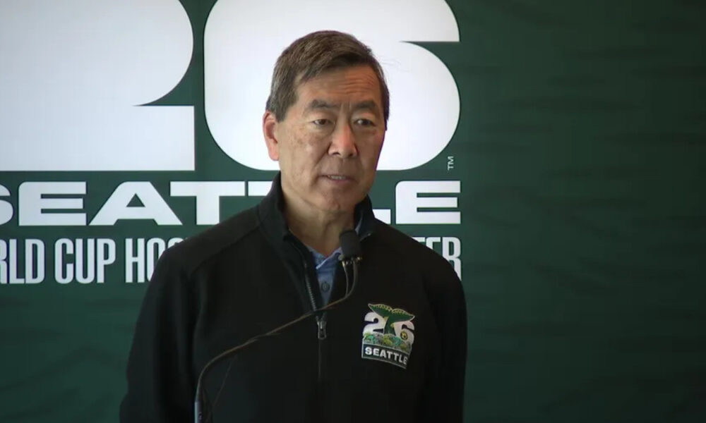 Man in a dark zippered jacket speaks into a microphone at a Seattle World Cup Hockey event, backdrop with large white lettering.