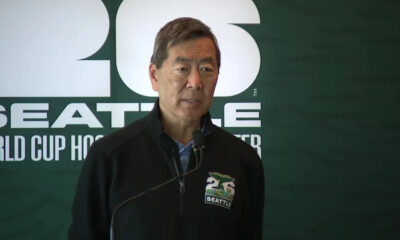 Man in a dark zippered jacket speaks into a microphone at a Seattle World Cup Hockey event, backdrop with large white lettering.