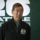 Man in a dark zippered jacket speaks into a microphone at a Seattle World Cup Hockey event, backdrop with large white lettering.