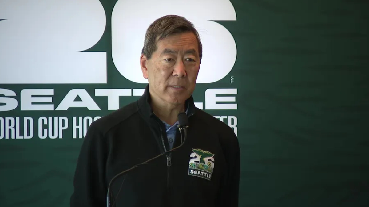 Man in a dark zippered jacket speaks into a microphone at a Seattle World Cup Hockey event, backdrop with large white lettering.